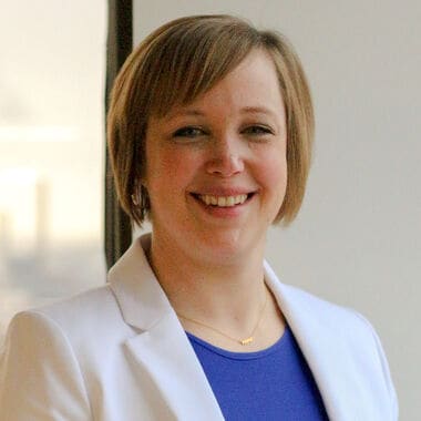A woman with short, blonde hair wearing a blue top with a white suit jacket.